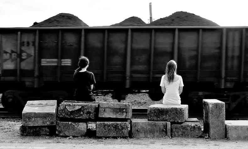 As artistas Bárbara Lissa e Maria Vaz sentadas em banco de estação de trem, vistas de costas 