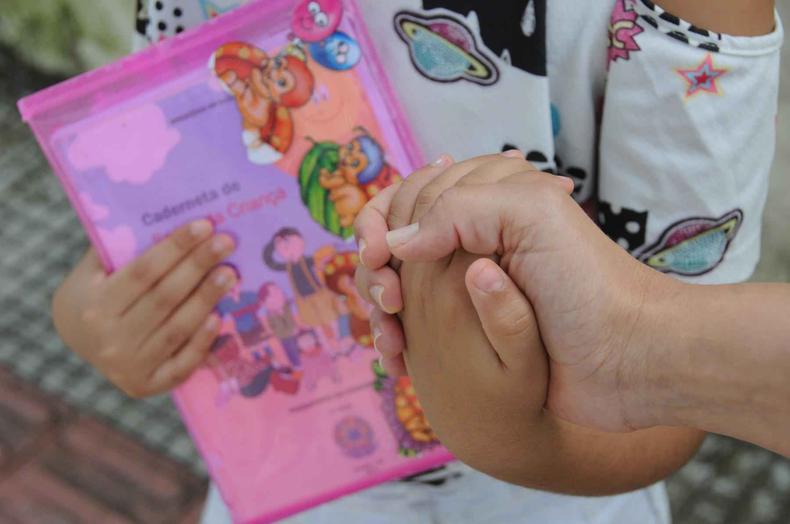 Detalhe da mo de uma me segurando a mo da filha com a caderneta de vacinao de cor rosa ao fundo