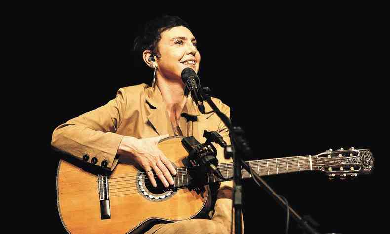 Sentada e segurando o violo, a cantora Adriana Calcanhotto sorri diante do microfone, durante um show