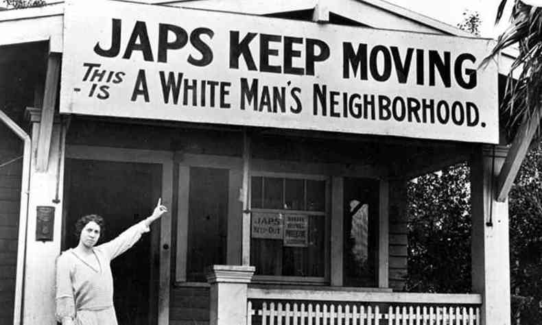 Mulher branca aponta para uma placa com os dizeres 'Japs keep moving, this is a white man's neighourhood', traduzido para 'Japs, continuem se mudando, este  um bairro de homens brancos'