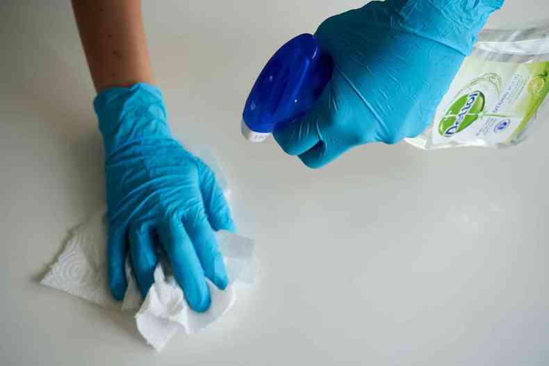Mos com luvas azuis limpando superfcie, uma segura um papel toalha e a outra um produto de limpeza.