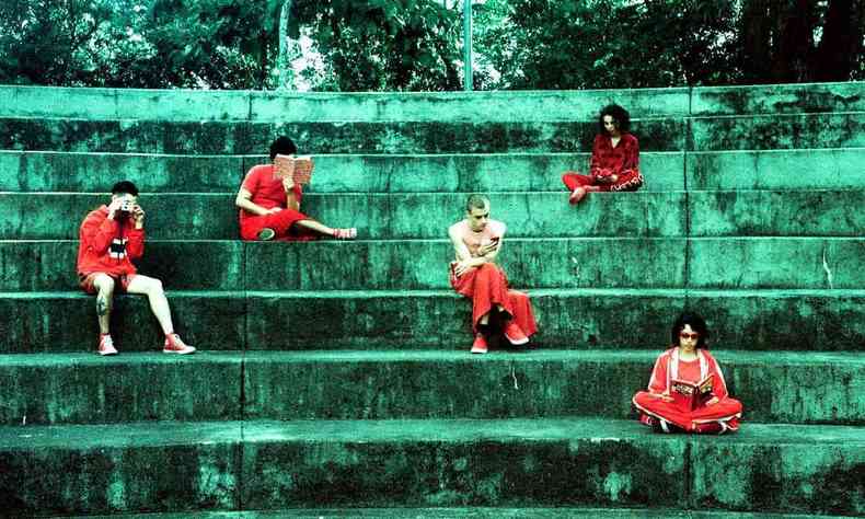  Em escada de cimento esverdeada, com rvores atrs, em formato de arena, cinco pessoas sentadas em diferentes degraus, vestidas de vermelho, mantm suas cabeas baixas, lendo um livro 