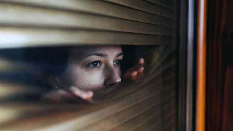 Mulher olhando pela fresta de persiana na janela