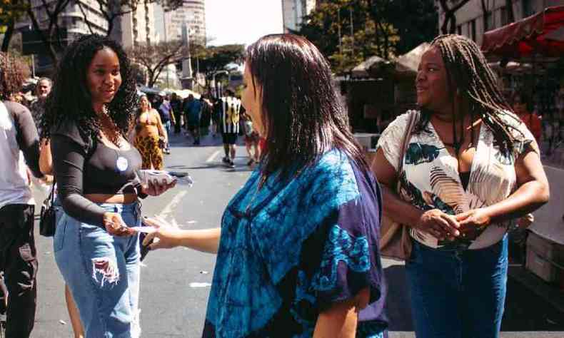 Jovem negra de cabelos longos e crespos entrega panfleto para duas mulheres na feira hippie