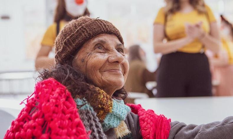 Mulher idosa de pele parda e cabelos castanhos cacheados usa uma touca de tric e uma echarpe tambm de tric. Ela sorri para algum alm da cmera