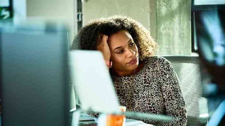 Mulher negra de cabelo encaracolado olha para o lado com olhar distante; ela est sentada em frente a um computador