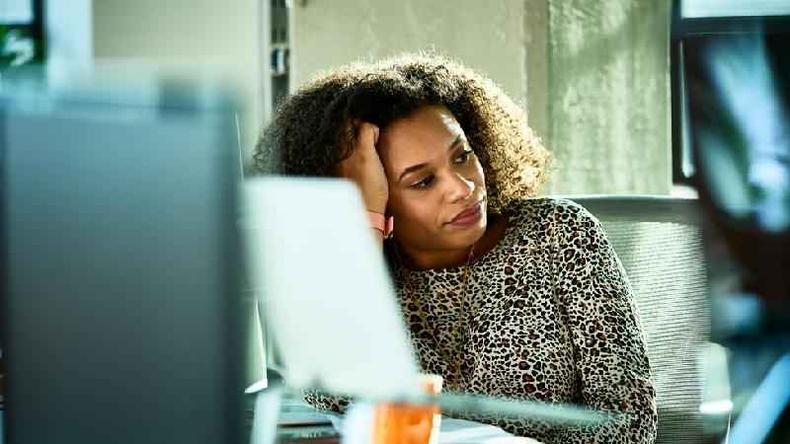 Mulher negra de cabelo encaracolado olha para o lado com olhar distante; ela est sentada em frente a um computador