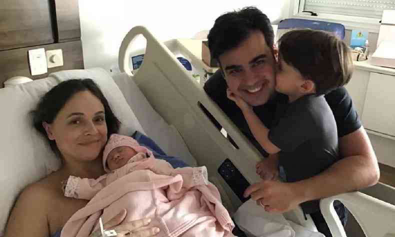 Mulher em cama de hospital com a filha recm nascida no colo, acompanhada do marido e filho de trs anos