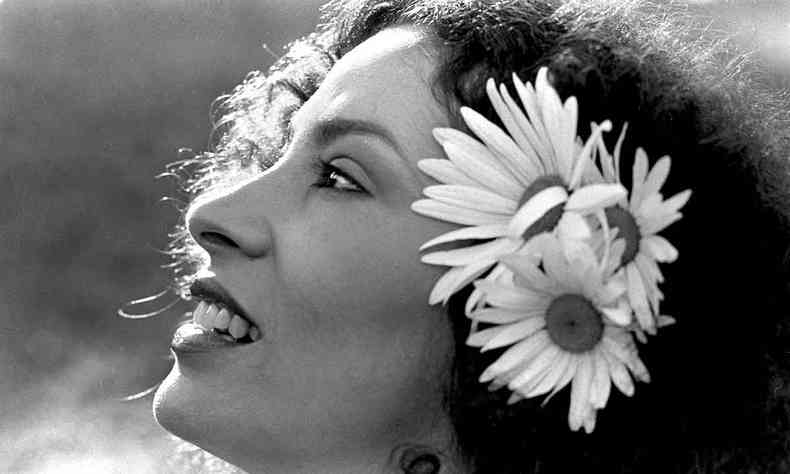 A cantora Clara Nunes sorri, de perfil, com flores no cabelo 