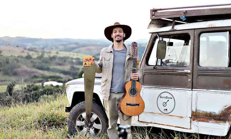 Usando roupa cinza, de p ao ar livre em frente a uma Rural, segurando um serrote e um cavaquinho, Guito sorri 