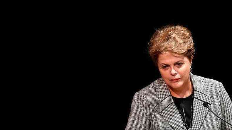 Dilma com olhar um pouco srio em p, diante de microfone e fundo preto