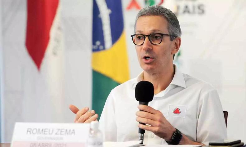 Romeu Zema sentado em uma mesa, com um microfone na mo, e as bandeiras de Minas Gerais e do Brasil ao fundo