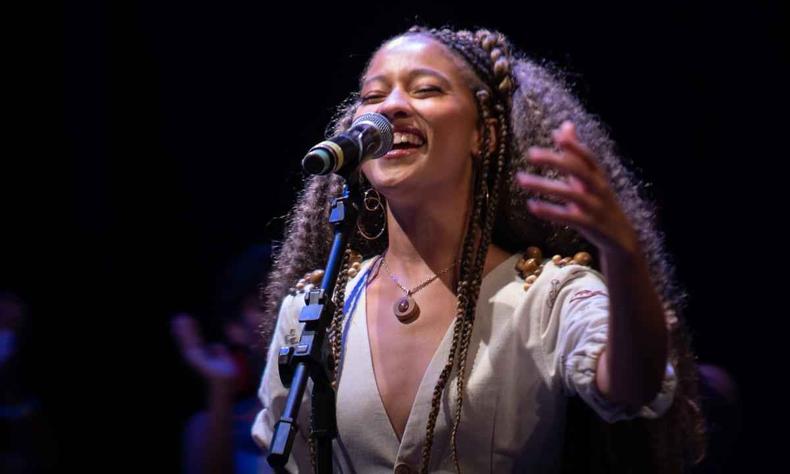 Vitria Pires, jovem negra com cabelos longos e caceados, cantando ao microfone em um fundo preto