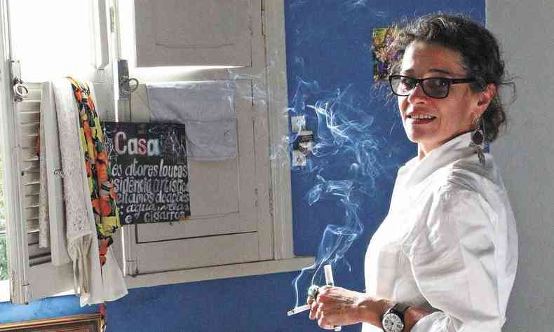 De calça jeans e camisa branca, com cigarro na mão, a atriz Viviane de Cassia Ferreira olha para a câmera 
