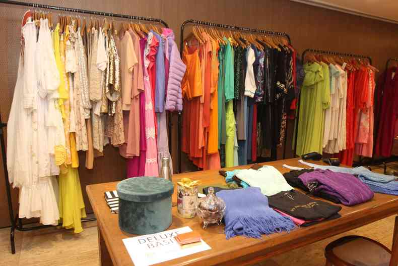 Roupas coloridas dispostas em uma arara no segundo plano, em primeiro plano uma mesa com diversas roupas dobradas. 