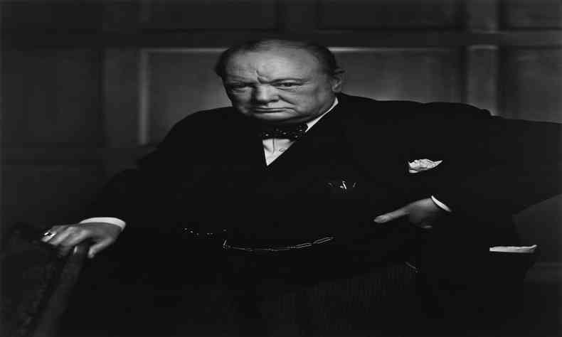 Imagem em preto e branco do primeiro ministro britnico Winston Churchill em p com uma das mos em uma cadeira