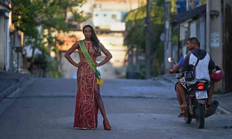 Elo Rodrigues est no meio da rua, com roupa de gala e a faixa de miss; uma moto passa com dois jovens que olha para ela