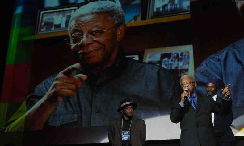 Milton Gonalves fala ao microfone no palco com imagem sua projetada em telo, atrs