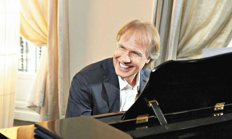 Vestindo terno preto e sentado ao piano, Richard Clayderman sorri 