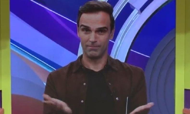 Tadeu Schmidt, um homem branco de cabelos castanhos, faz gesto com as m�os em Libras. Ele usa uma camiseta preta e uma camisa marrom