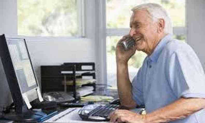 Homem branco de cabelos grisalhos fala ao telefone e mexe em um computador. Ele usa uma camisa social azul clara de mangas curtas