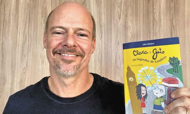 Léo Dicker sorri e mostra para a câmera a capa do livro Clara e Guto - Os segredos de Londres