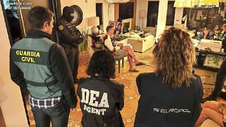 Fotografia colorida mostra policiais de p em um apartamento cheio de documentos e pessoas