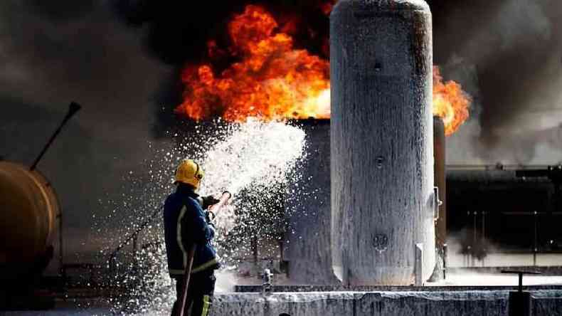 Fotografia colorida mostra bombeiro usando mangueira para apagar fogo em um imvel que parece ser industrial