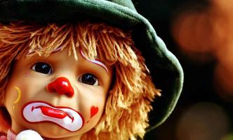 Boneco palhao com chapu e cabelo volumoso feito de linhas de l, rosto pintado de branco ao redor da boca e corao desenhado na bochecha e nariz pintado de vermelho.