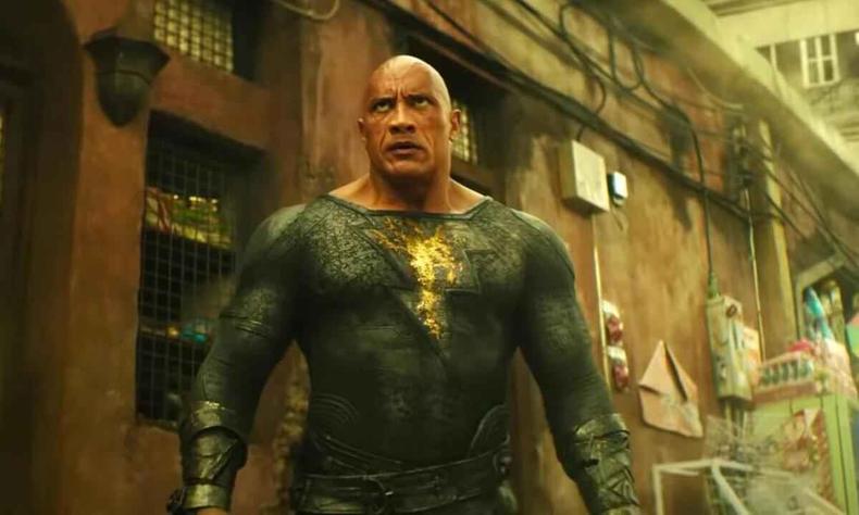 Em solo de areia, o ator Dwayne Johnson, agachado, segura garoto em cena de Ado Negro