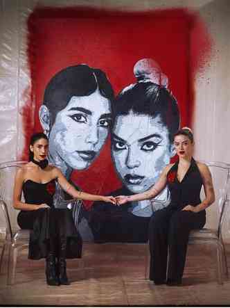 Sentadas, as cantoras Clara e Sofia do as mos na capa de seu disco, tendo ao fundo pintura dos respectivos rostos sob fundo vermelho