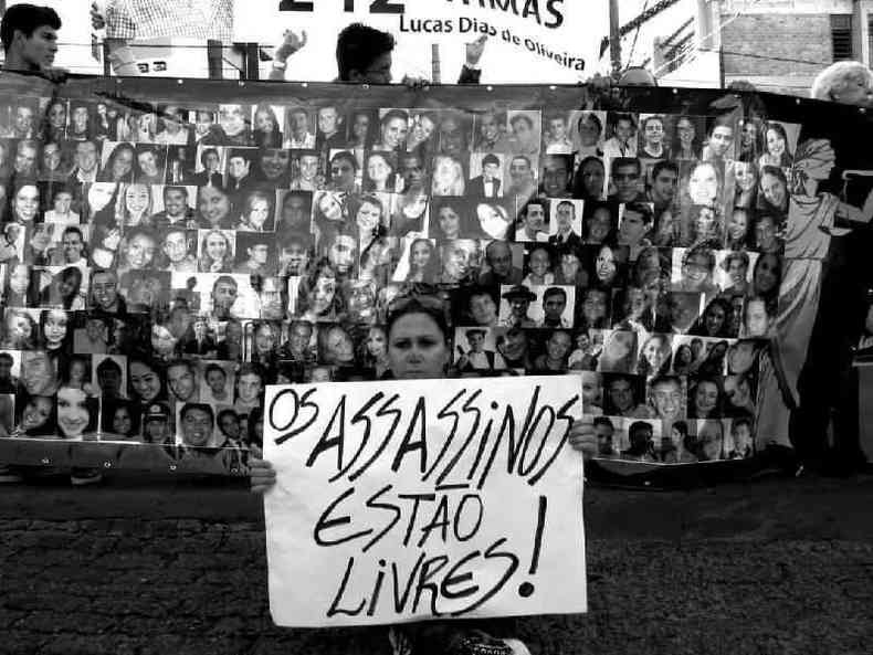 Mulher em frente a mural com fotos das vtimas segurando cartaz em que se l: 'Os assassinos esto livres'
