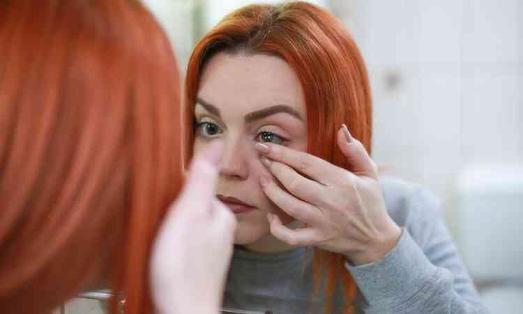Mulher de cabelo ruiva se prepara para colocar a lente de contato diante de um espelho