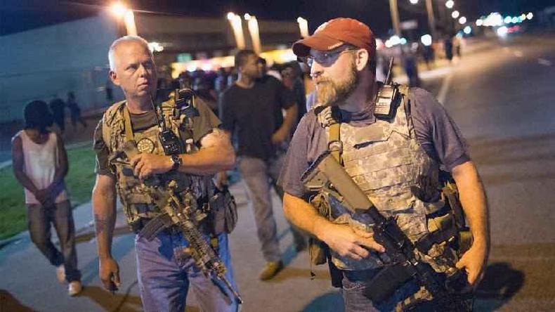 Fotografia mostra vrias pessoas armadas, em primeiro plano, dois homens com coletes e armas