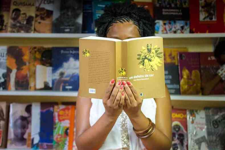 Uma mulher negra de cabelo preto tranado segura um livro em frente ao seu rosto. Ao fundo, uma estante cheia de livros