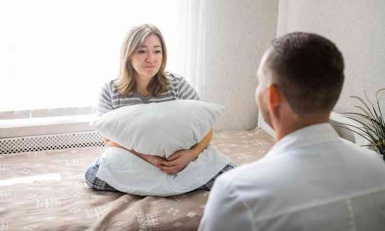 mulher de pernas cruzadas em cima da cama e travesseiro no colo olhando para um homem sentado a sua frente