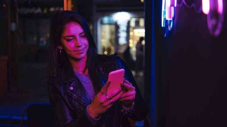 Mulher branca de cabelo preto no ombro com piercing no nariz olha para a tela de um celular; ela est uma boate e  iluminada por uma luz rosa