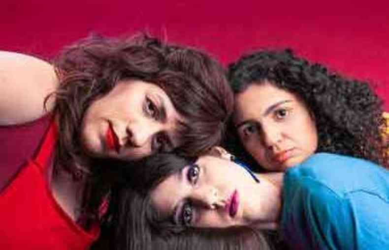 Em cenrio de fundo vermelho, Julia Branco, Luiza Brina e Sara No Tem Nome amontoam suas cabeas em cima de uma mesa 