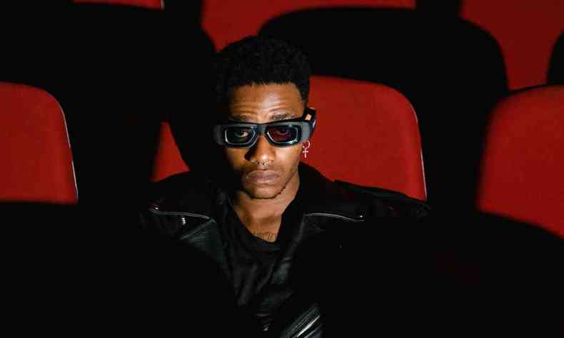 Homem negro, com jaqueta preta e culos 3D, sentado sozinho em uma sala de cinema com poltronas vermelhas