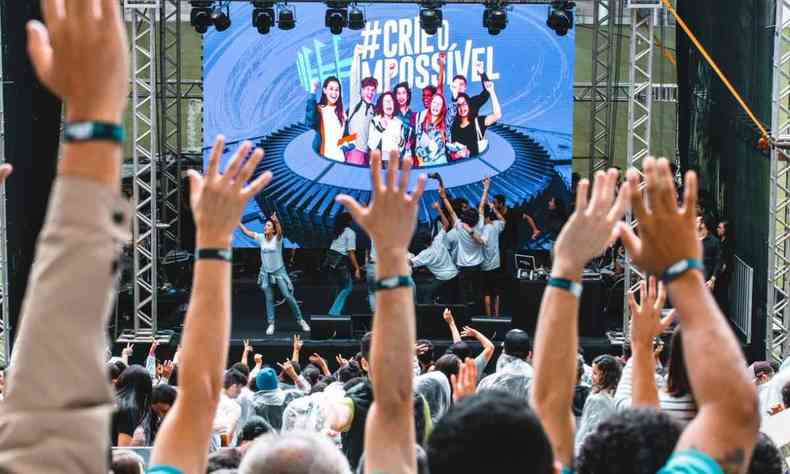 Jovens com as mãos levantas assistem pessoas em um palco do evento
