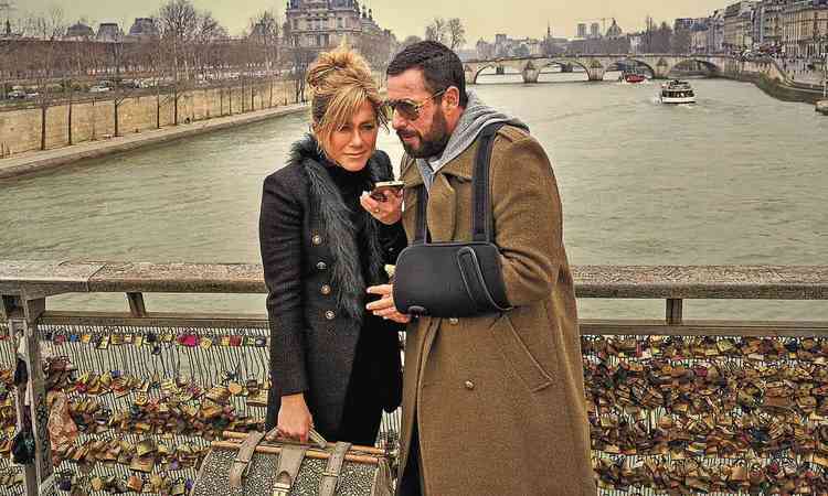 Atores Jennifer Aniston e Adam Sandler, vestindo roupas de frio, olham no celular. Ao fundo v-se o Rio Sena, no filme 'Mistrio em Paris'
