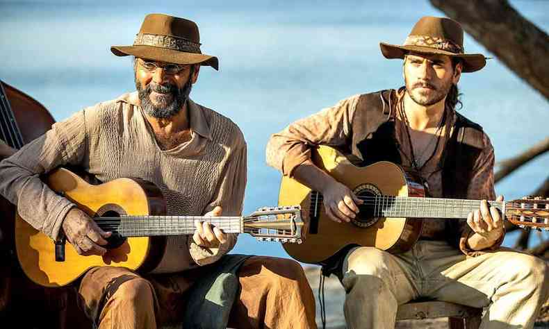 Almir e Gabriel Sater, sentados, tocando violo e sorrindo, em cena da novela Pantanal 