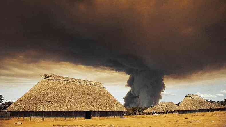 Fotografia colorida mostra trs construes de uma aldeia com uma coluna de fumaa preta ao fundo