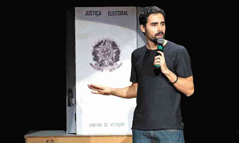 O humorista Bruno Costoli aparece em cena, segurando o microfone, em frente a rplica de uma cabine de votao