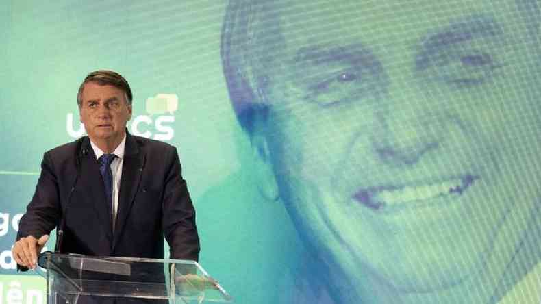 Bolsonaro com feio sria falando no microfone, com foto dele sorrindo atrs em painel