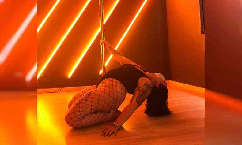 Ravena Carvalho uma mulher gorda, faz uma pose sensual no cho se apoiando na barra de pole dance em um quarto escuro com luzes vermelhas