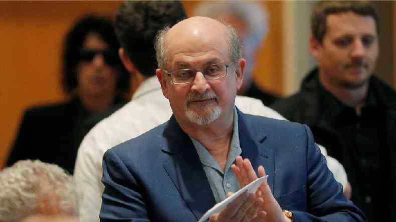 Salman Rushdie olha para a cmera em primeiro plano, com pessoas ao fundo