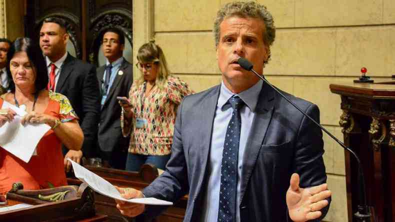 Leonel Brizola Neto na Cmara Municipal do Rio de Janeiro. A foto o enquadra da cintura para cima. Ele veste um terno cinza, uma camisa azul-claro e uma gravada estampada em cinza e azul. O poltico fala com microfone e segura um papel. Ao fundo, h duas mulheres e dois homens. 