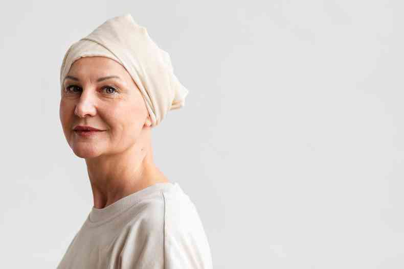 Mulher que passou pelo câncer com um turbante branco na cabeça. Ela usa blusa branca e está sorrindo 