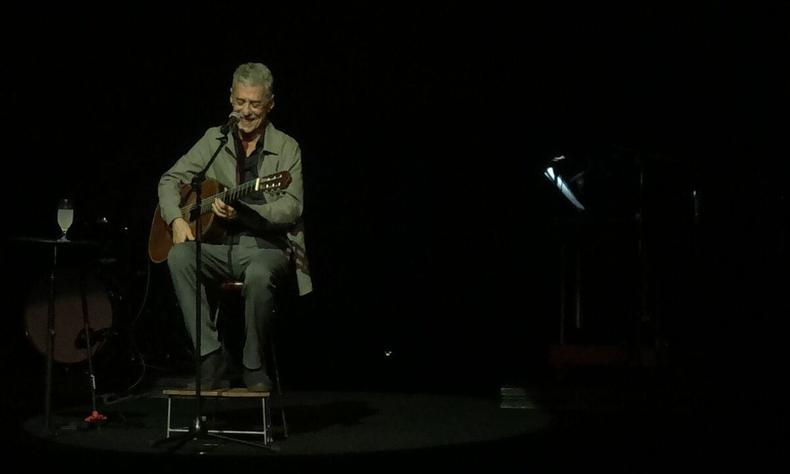 Com roupas de tom cinza, sentado no banquinho com um violo, Chico Buarque sorri
